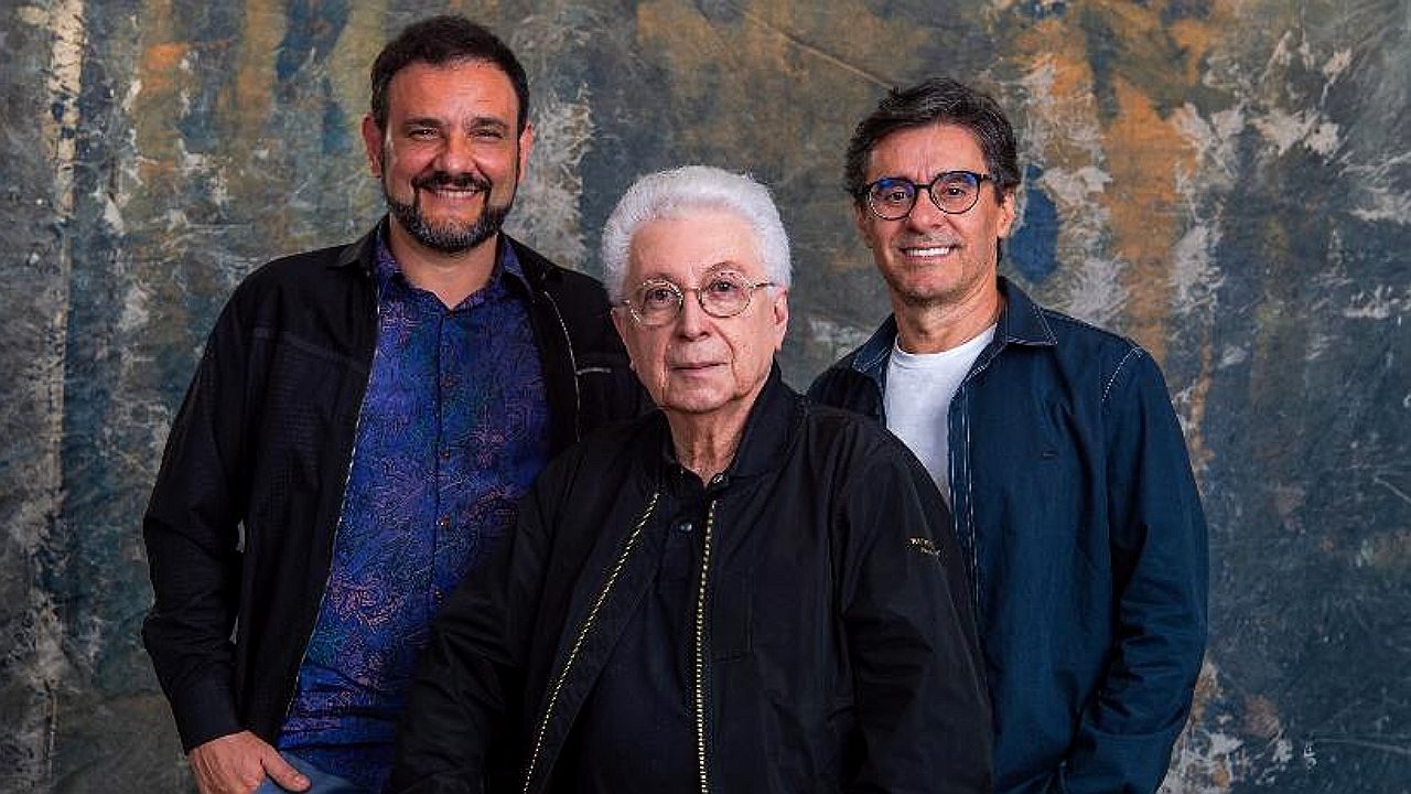Três homens posam juntos para foto em estúdio com fundo artístico texturizado