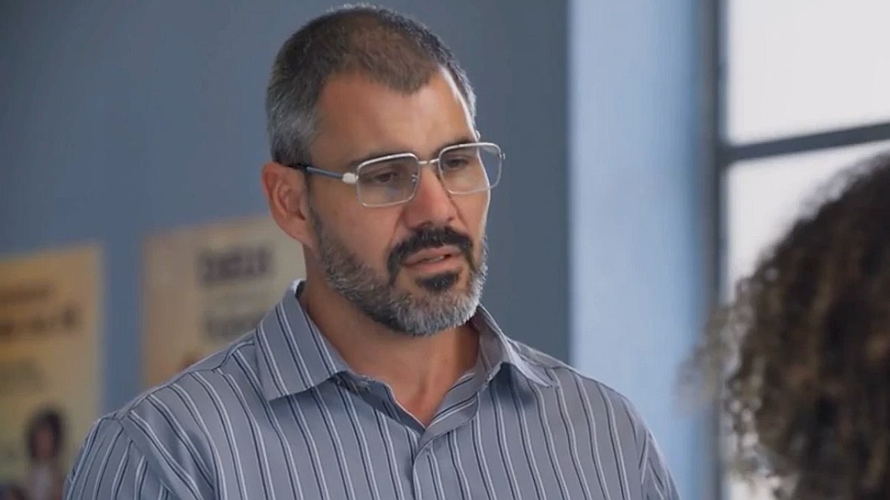 Homem de óculos e barba conversa com outra pessoa em ambiente interno em cena de novela