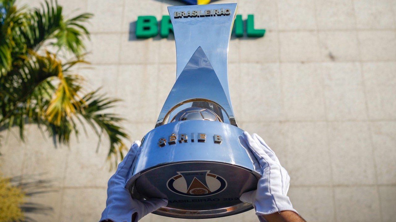Troféu da Série B do Brasileirão erguido por mãos com luvas brancas em frente à sede da CBF com nome Brasil ao fundo