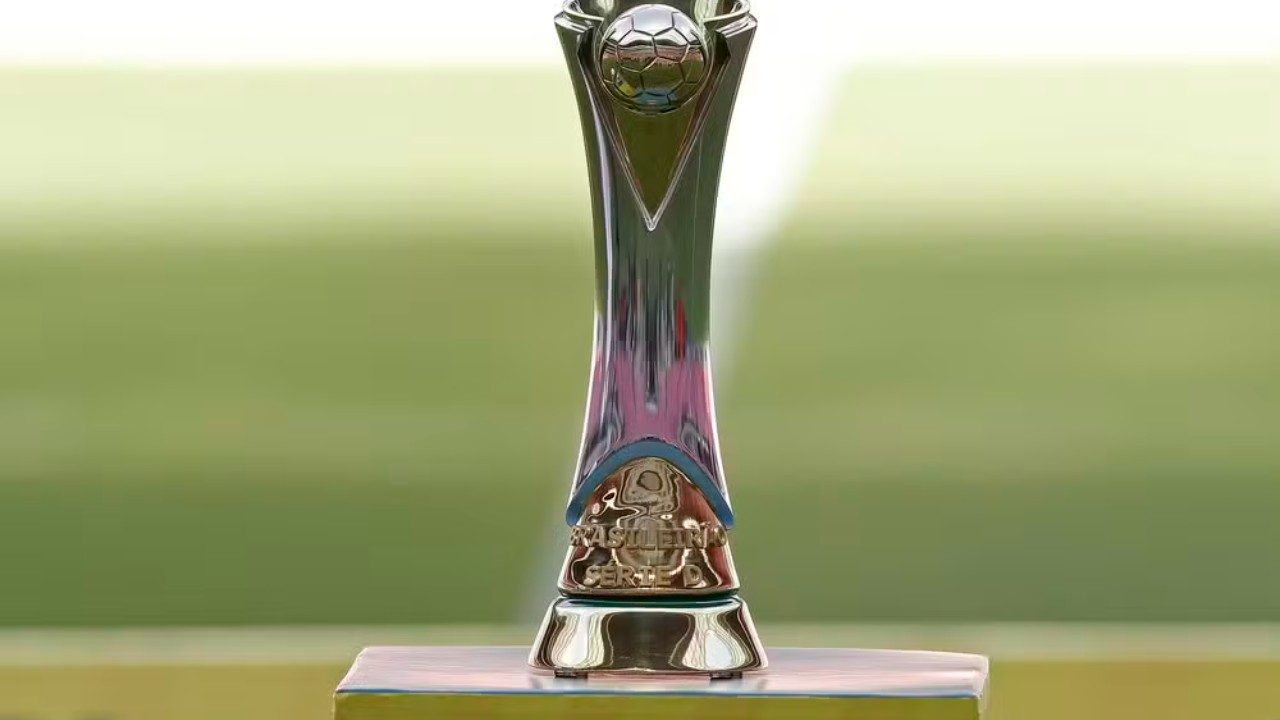 Troféu de prata do Brasileirão Série D com bola de futebol no topo posicionado sobre pedestal de madeira, diante de gramado verde desfocado ao fundo