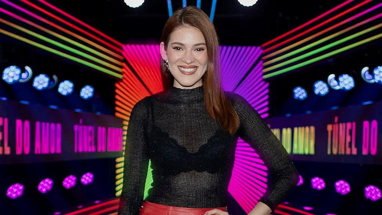 Mulher ruiva posa sorrindo em cenário colorido com luzes neon do programa Túnel do Amor