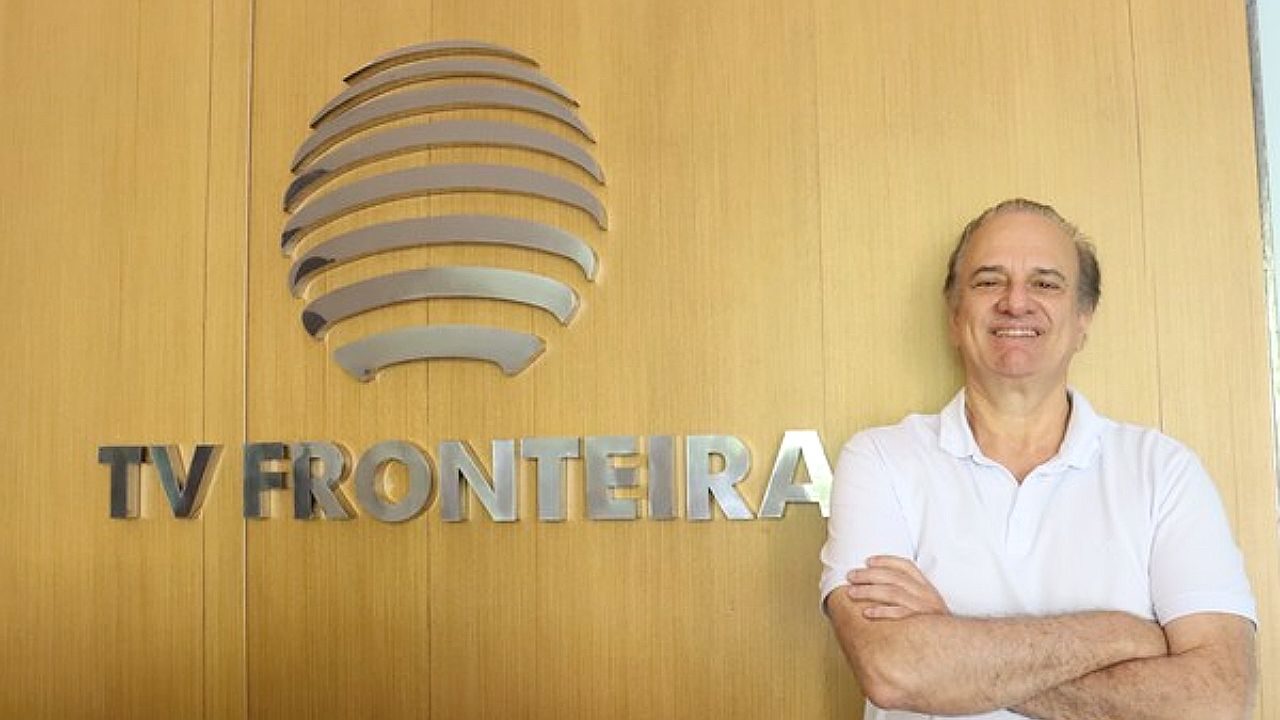 Homem sorri com braços cruzados diante de parede com logotipo da TV Fronteira em ambiente corporativo