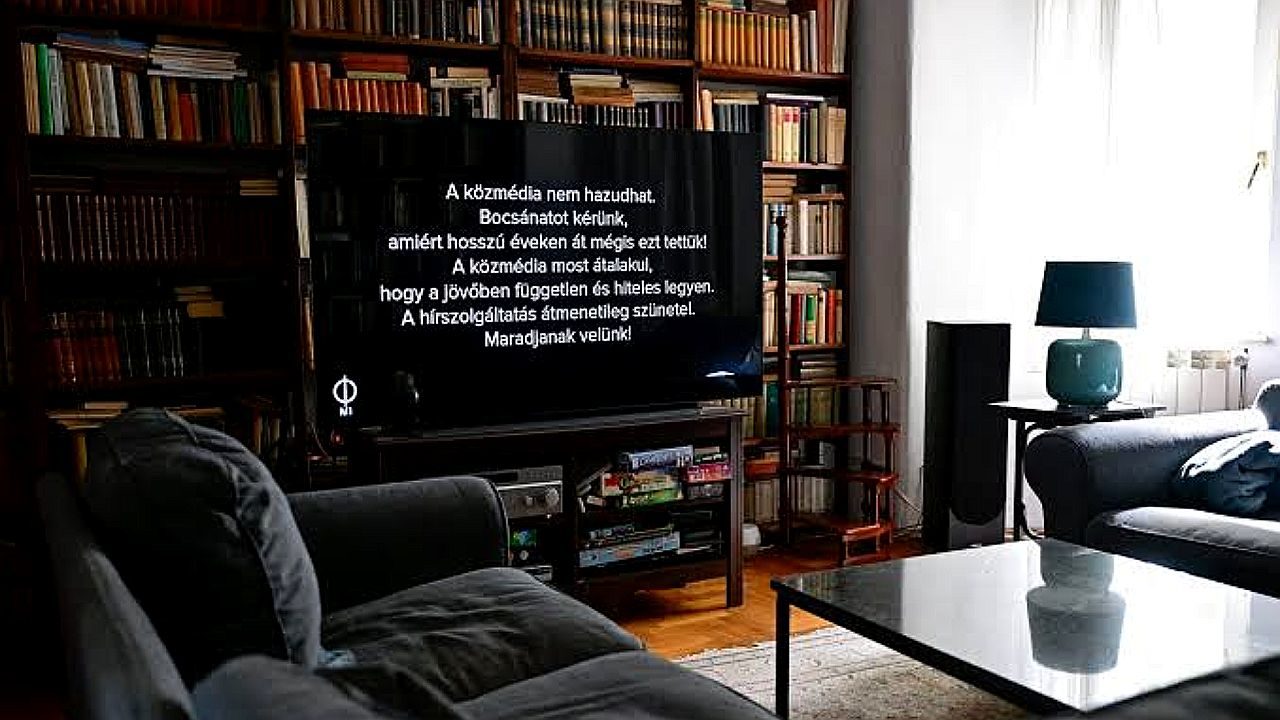 Tela de TV exibe aviso de interrupção da programação da emissora pública em sala de estar com estante de livros ao fundo