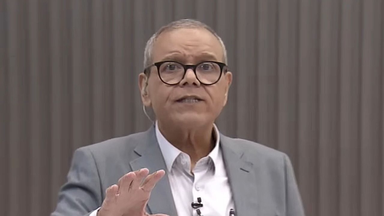 Apresentador de óculos e terno cinza gesticula durante fala em estúdio de TV com fundo neutro vertical