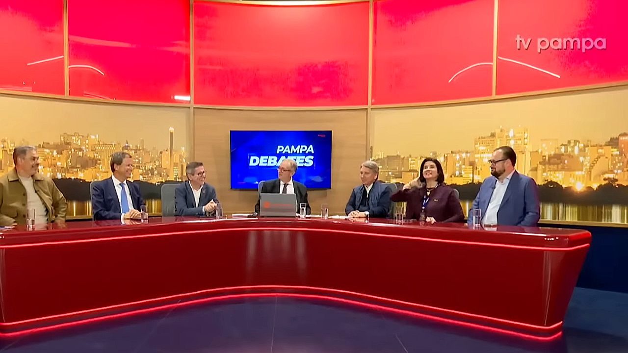 Participantes sentados em bancada curva vermelha durante edição do programa Pampa Debates em estúdio da TV Pampa com telão ao centro e cenário urbano ao fundo