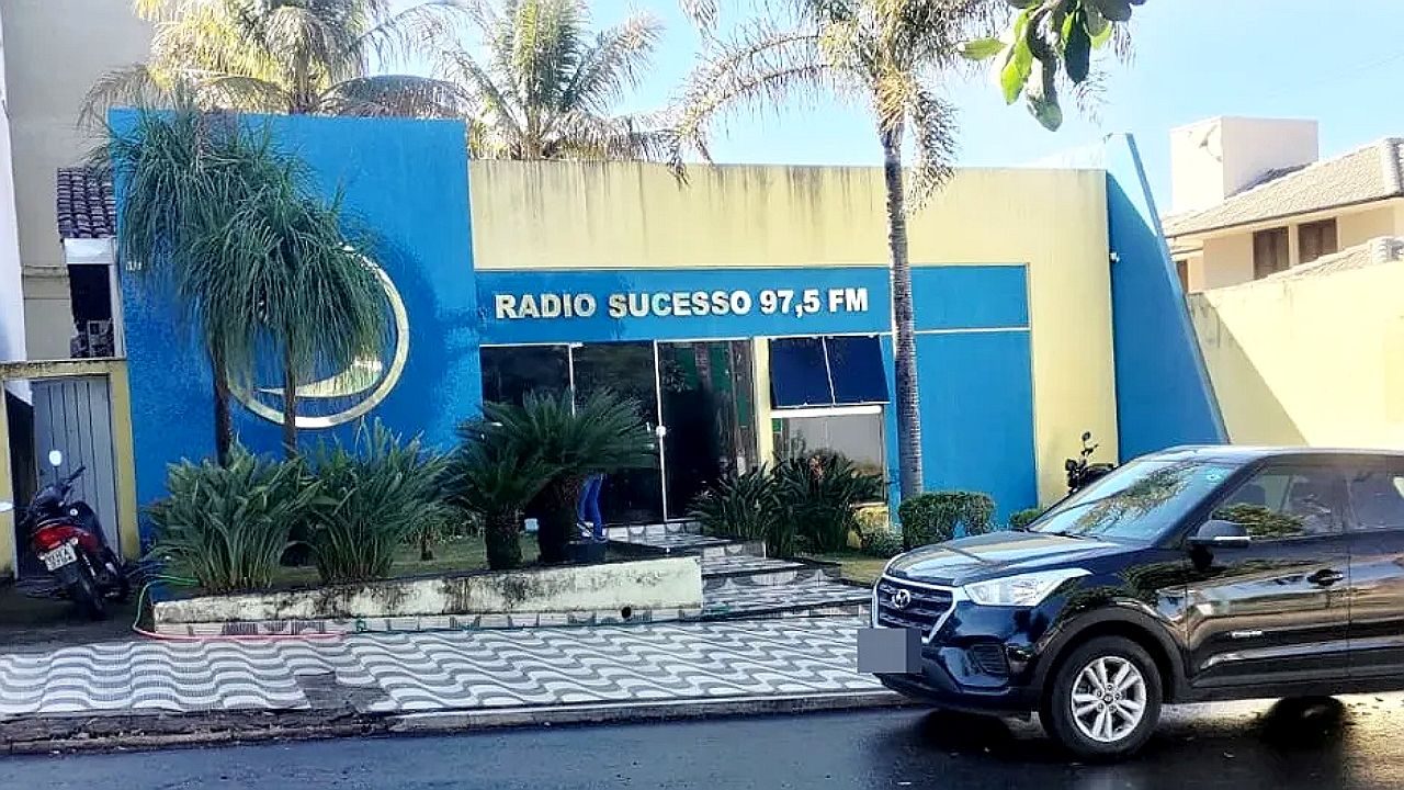 Fachada da Rádio Sucesso 97,5 FM com prédio azul e amarelo e carro estacionado em frente