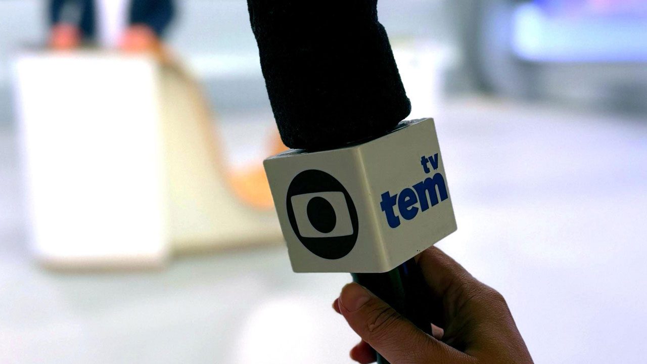 Microfone com logo da Globo e identificação da afiliada TV Tem sendo segurado por uma mão em estúdio de TV