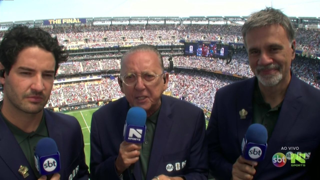 Galvão Bueno, com microfone entre dois comentaristas, em cabine com estádio lotado ao fundo na final da Copa do Mundo