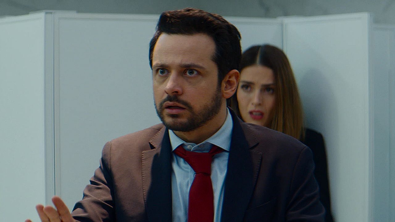 Homem de terno e gravata vermelha demonstra preocupação enquanto mulher observa ao fundo em cena de novela mexicana