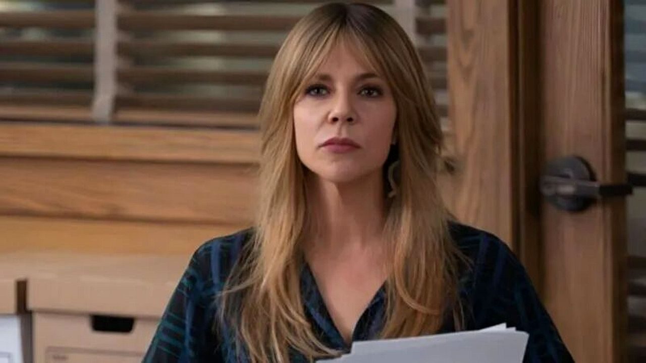 Mulher loira segura folhas de papel e encara à frente com expressão séria em ambiente interno com móveis de madeira e persianas ao fundo, sugerindo cena de escritório ou reunião.