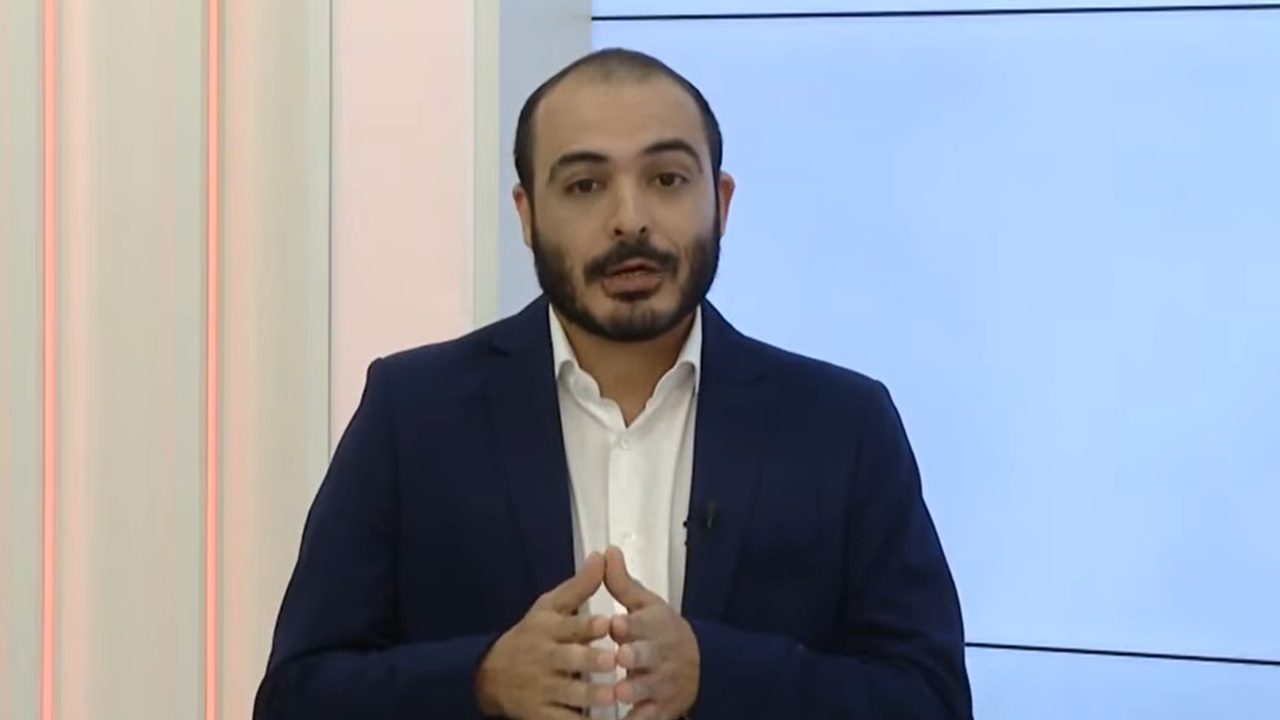 Homem apresenta programa de TV em estúdio iluminado, usando terno azul-escuro e camisa branca, com as mãos unidas à frente do corpo.