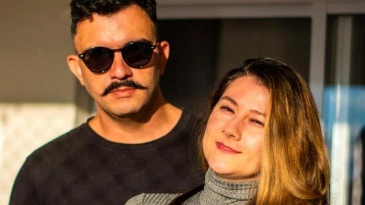 Vagner Macedo de óculos escuros ao lado de Bella Mantovani de blusa cinza diante de porta de vidro sob luz do sol
