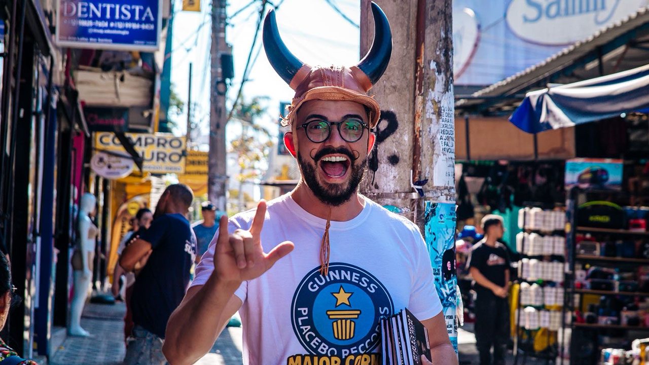 Vagner Macedo sorri usando chapéu com chifres e camiseta branca em rua de comércio popular