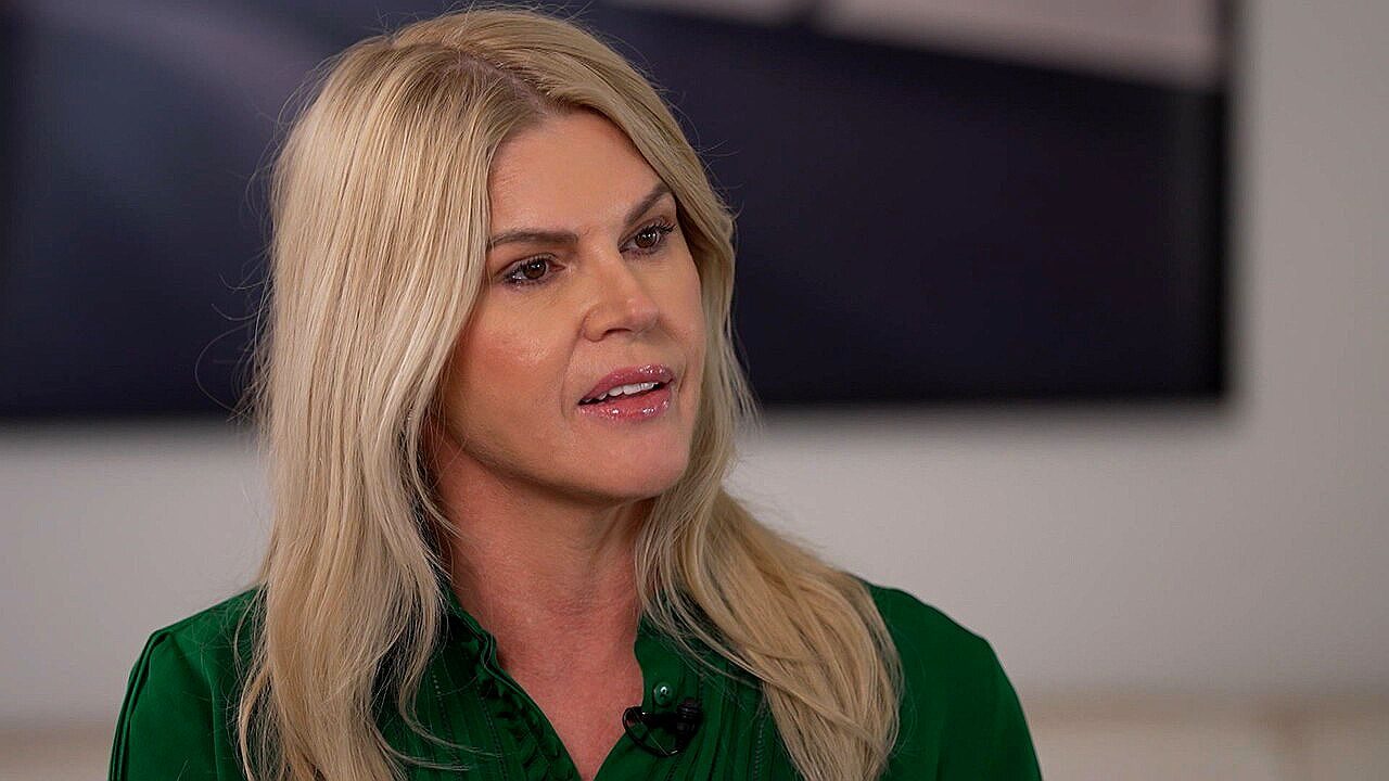 Mulher loira de cabelos longos conversa durante entrevista em ambiente interno, usando blusa verde e microfone de lapela, com expressão atenta enquanto olha para alguém fora de quadro