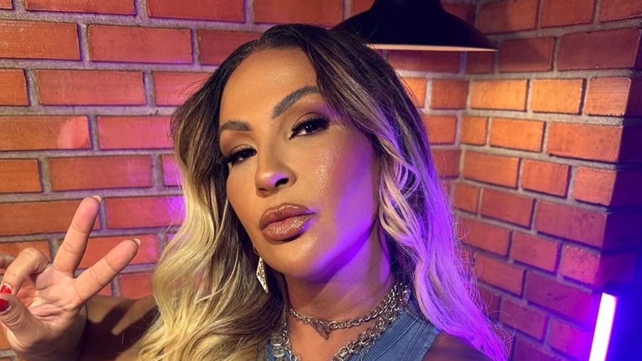 Mulher adulta posa para a câmera em ambiente interno com parede de tijolos aparentes. Ela tem cabelos longos com mechas claras, usa maquiagem marcante e acessórios metálicos no pescoço e nas orelhas. Com expressão confiante, faz um gesto de paz com a mão. A iluminação colorida em tons de roxo e rosa cria um clima de estúdio ou bastidores de gravação.