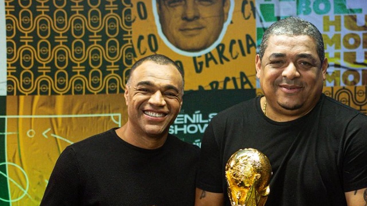 Dois homens sorriem lado a lado diante de um painel decorado com imagens e referências esportivas. Ambos vestem camisetas pretas; o homem à direita segura um troféu dourado em formato de taça. O registro sugere participação em programa ou evento ligado ao futebol.