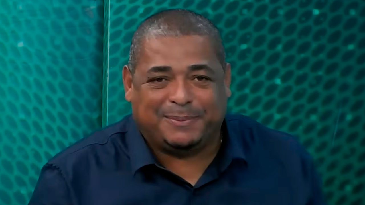 Ex-jogador Vampeta sorri para a câmera vestindo camisa social azul escura, posicionado em frente a um painel verde com textura de pequenos círculos