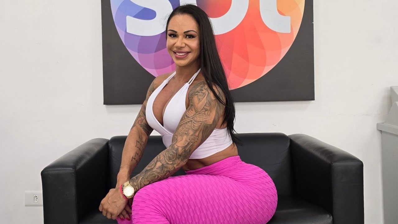 Vanessa Ataídes sorridente sentada em sofá preto usa top branco e calça rosa diante do logotipo colorido do SBT