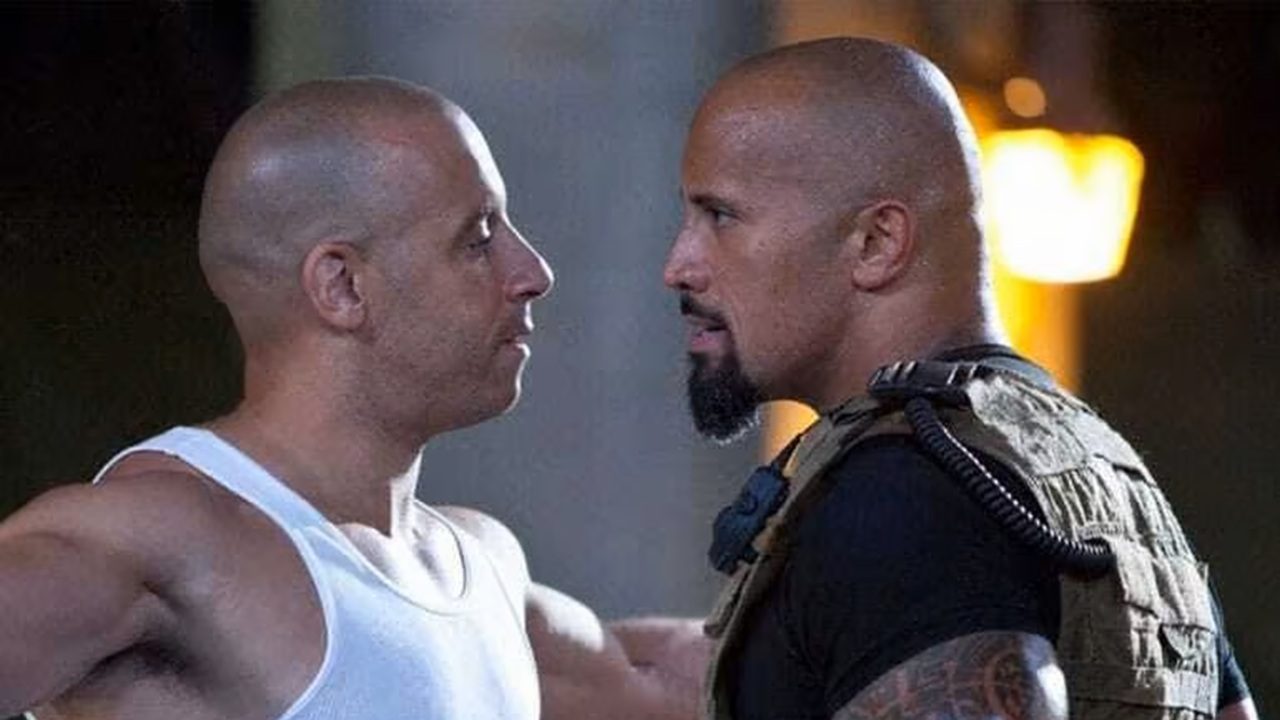 Dois homens carecas e musculosos aparecem frente a frente, muito próximos, em um confronto tenso. O homem à esquerda usa uma camiseta branca sem mangas; o da direita veste roupa escura com um colete tático e tem barba curta. Ambos mantêm expressões sérias e olhares fixos um no outro. O fundo é escuro, com iluminação quente desfocada, sugerindo uma cena de ação ou conflito intenso em um filme.