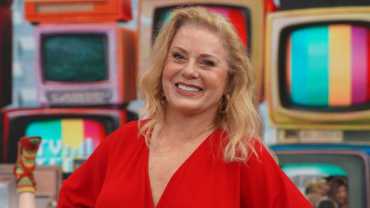 Vera Fischer sorrindo com vestido vermelho no palco do Caldeirão com Mion diante de cenário com televisores coloridos