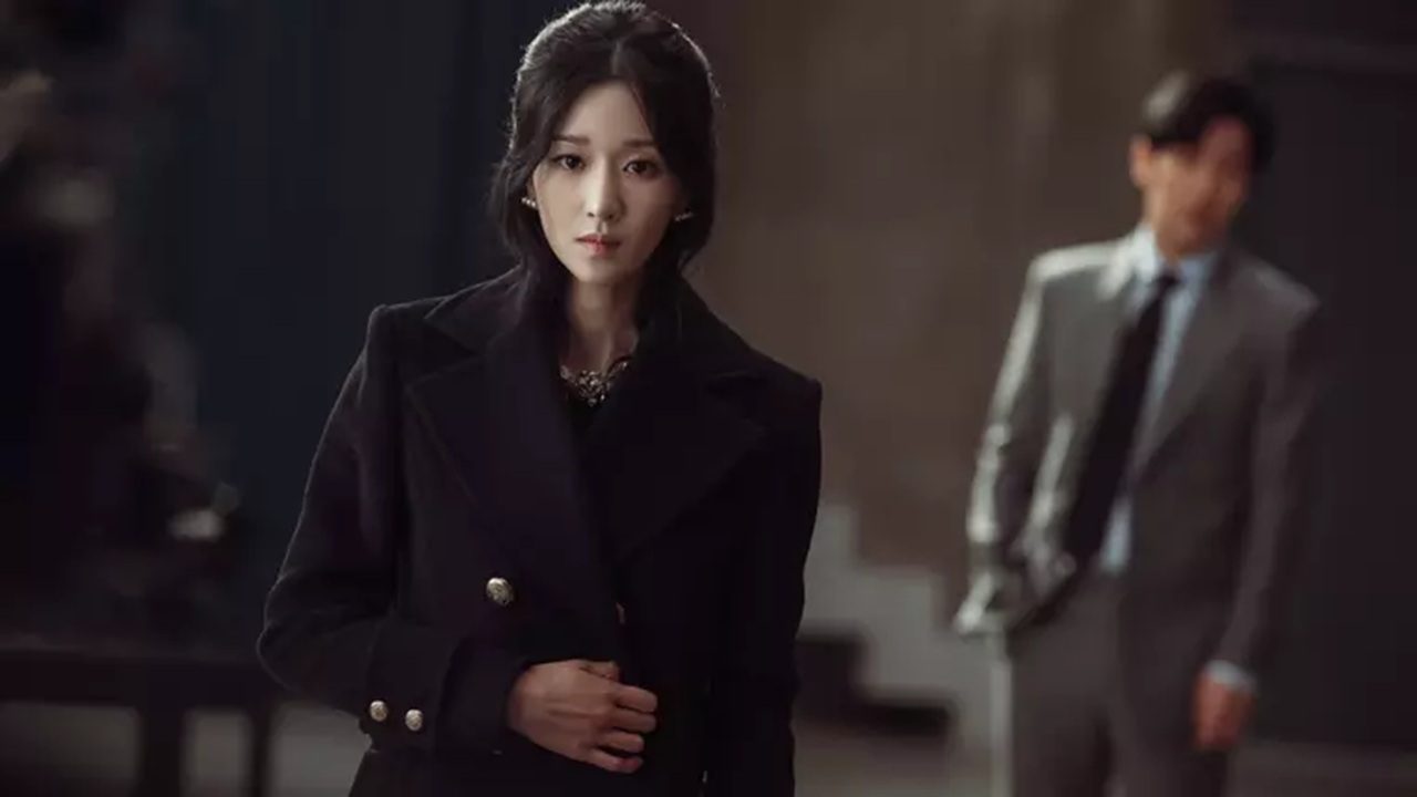 Mulher de casaco preto encara à frente enquanto homem de terno desfocado aparece ao fundo em ambiente interno elegante