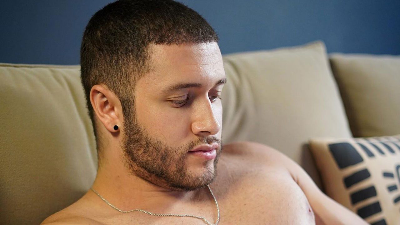 Victor Ferraz sem camisa sentado no sofá com expressão séria e olhar voltado para baixo