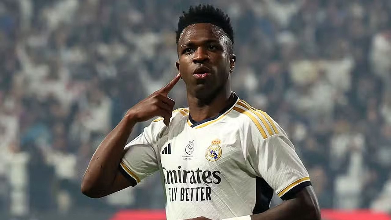 Vinícius Júnior aponta para o ouvido durante comemoração em campo usando uniforme do Real Madrid diante de arquibancada lotada desfocada ao fundo