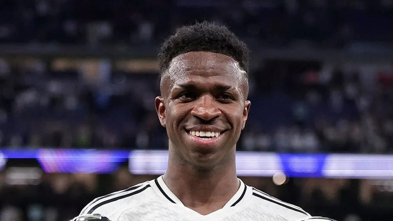Homem adulto negro em close frontal com expressão alegre e sorriso aberto mostrando os dentes, pele suada sugerindo atividade física recente, cabelo curto com laterais baixas e topo levemente mais alto, usa camisa esportiva branca com detalhes escuros nos ombros, olha diretamente para a câmera, ao fundo aparece um estádio de futebol desfocado com arquibancadas escuras e faixa horizontal iluminada em tons de azul e roxo, iluminação forte de estádio destaca o rosto e transmite clima de pós-jogo ou apresentação esportiva