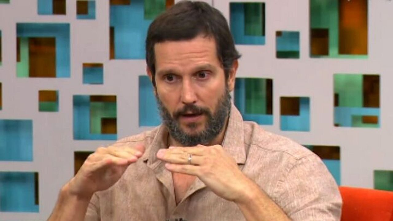 Homem de barba escura e cabelo curto participa de programa de TV, usando camisa bege de manga curta. Ele faz um gesto com as mãos na altura do peito, como se explicasse algo, diante de painel com recortes geométricos coloridos ao fundo.