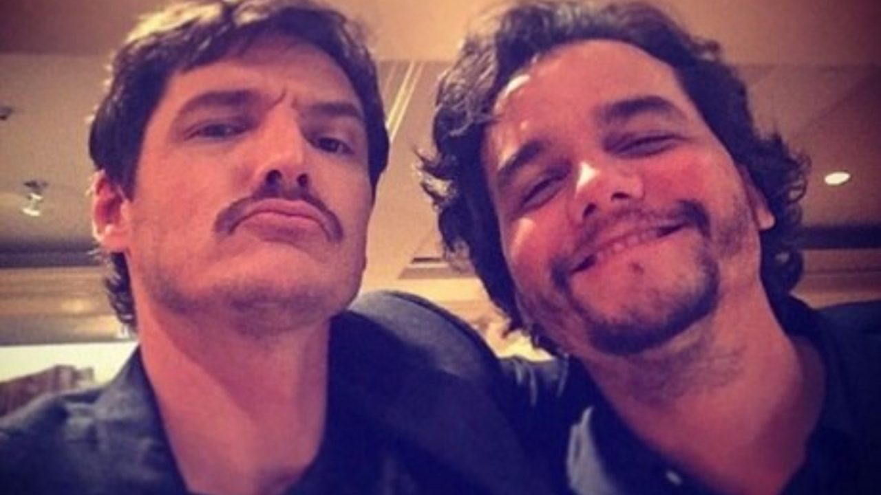 Dois homens barbados posam juntos sorrindo para selfie em ambiente interno com iluminação amarelada durante encontro informal