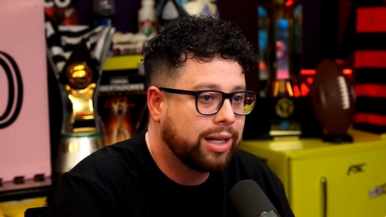 Homem jovem com cabelo curto e cacheado e barba aparada fala ao microfone em estúdio de podcast. Ele usa óculos de armação preta e camiseta escura. Ao fundo, há prateleiras com troféus, bola de futebol americano e objetos decorativos, em ambiente iluminado com luzes coloridas.