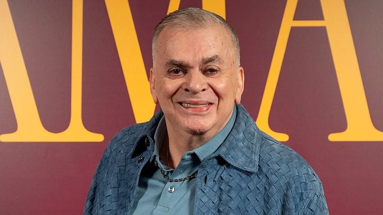 Homem de camisa azul posa sorrindo diante de painel com letras douradas ao fundo