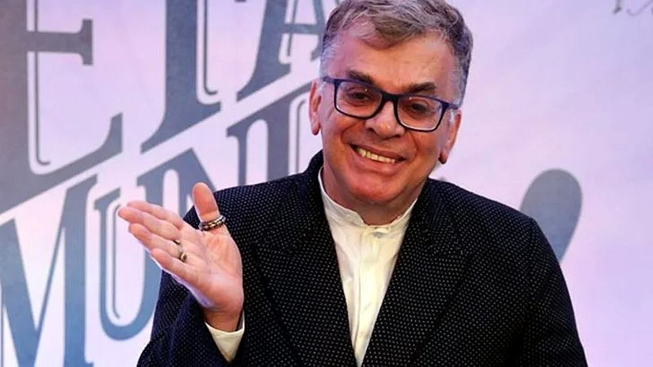 Um homem de meia-idade aparece sorrindo, usando óculos de armação escura e cabelo curto grisalho. Ele veste um blazer escuro com textura e uma camisa clara por baixo. Está com a mão levantada em gesto descontraído, exibindo alguns anéis nos dedos. O fundo é claro, com letras grandes desfocadas, sugerindo um cenário de evento, programa ou painel.