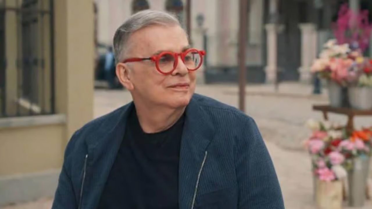 O autor de novelas Walcyr Carrasco, de cabelo grisalho e óculos vermelhos, posa sorridente em ambiente urbano ao ar livre. Ele veste camiseta preta e jaqueta azul, com vasos de flores coloridas ao fundo, transmitindo um clima leve e descontraído
