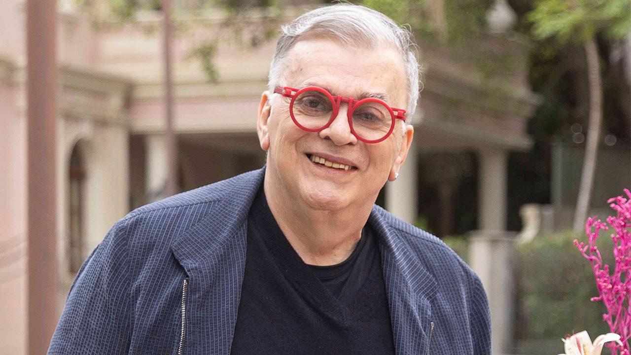 Walcyr Carrasco sorrindo com óculos vermelhos e blazer azul em ambiente externo com vegetação ao fundo