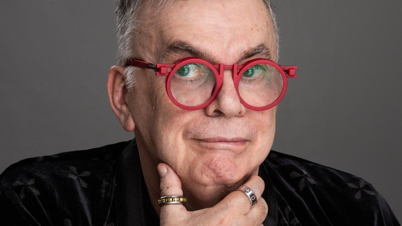 Walcyr Carrasco posa com óculos redondos vermelhos apoiando o queixo na mão em retrato de estúdio com fundo neutro