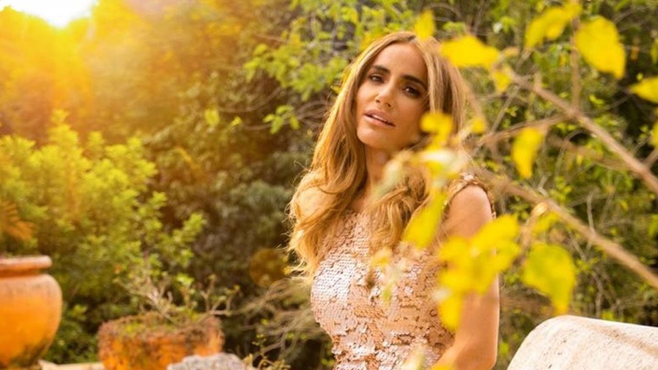 Wanessa Moura com vestido claro sentada ao ar livre cercada por vegetação e luz do pôr do sol ao fundo