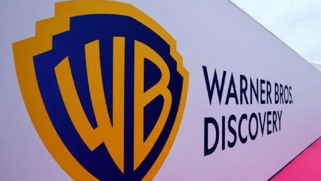 A imagem mostra o logotipo da Warner Bros. Discovery em um painel branco. À esquerda, está o icônico escudo dourado e azul com as iniciais “WB”, e à direita, o nome “Warner Bros. Discovery” aparece em letras maiúsculas pretas