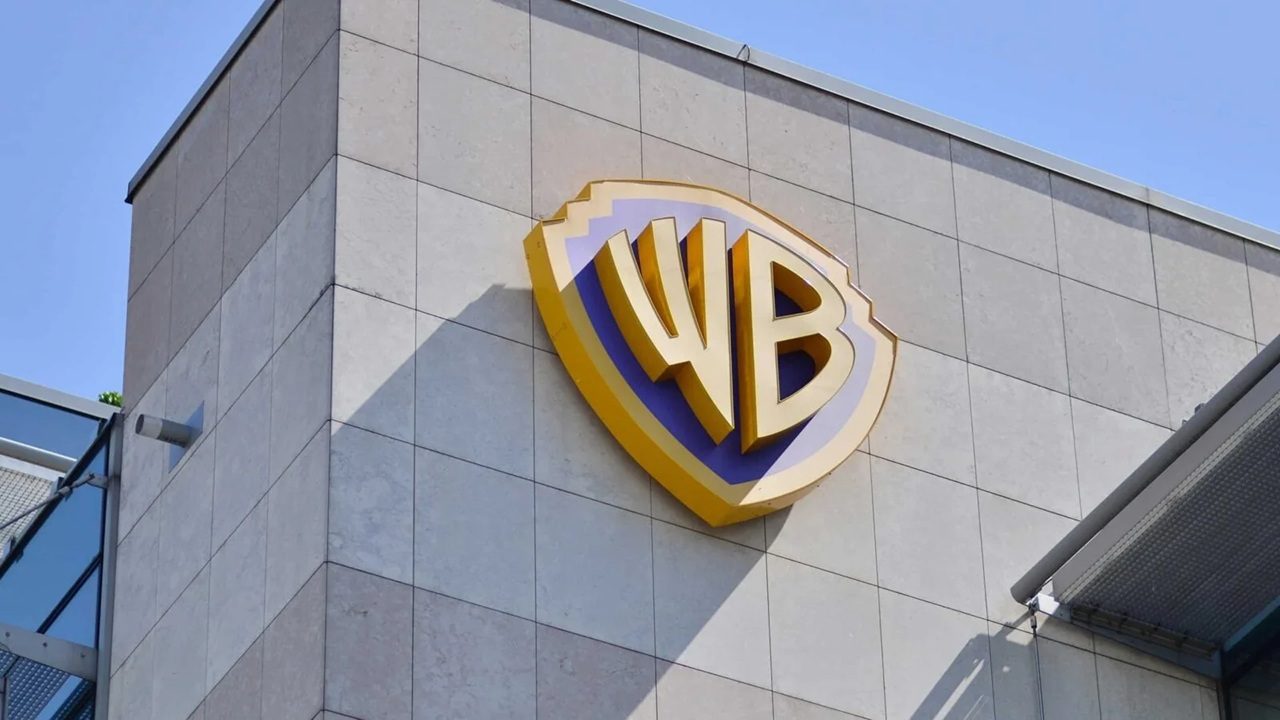 Logotipo dourado e roxo com as letras “WB” em relevo está fixado na parede externa de um prédio de fachada revestida por placas de pedra clara. A estrutura tem linhas modernas e, ao fundo, o céu está limpo e azul.