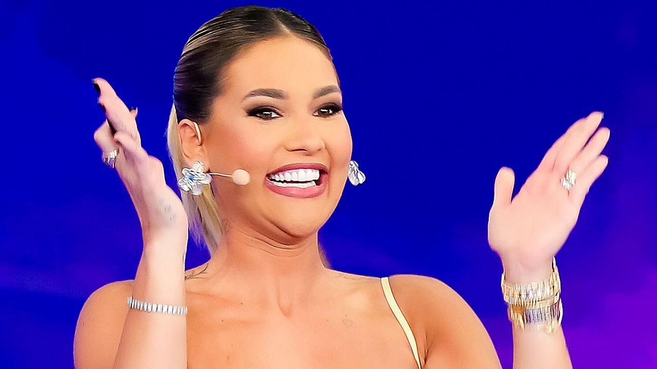 Mulher sorridente em estúdio de TV usa microfone de headset e joias brilhantes enquanto levanta as mãos, aparentemente apresentando ou reagindo a algo diante de um fundo azul iluminado.