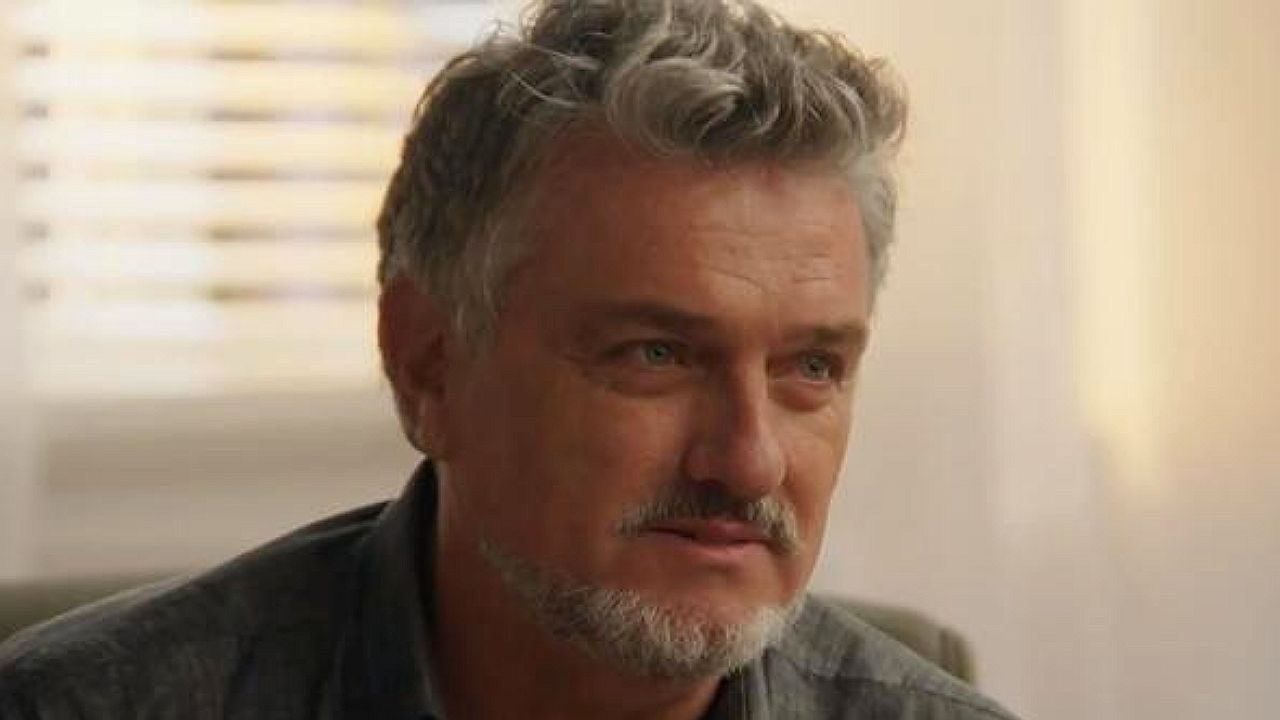 Homem grisalho com barba olha de lado em ambiente interno em cena de série dramática