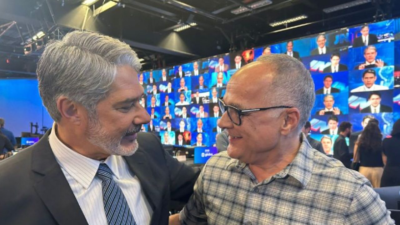 Dois homens se cumprimentam sorrindo em estúdio de televisão, com grande painel de telas exibindo imagens de telejornais ao fundo