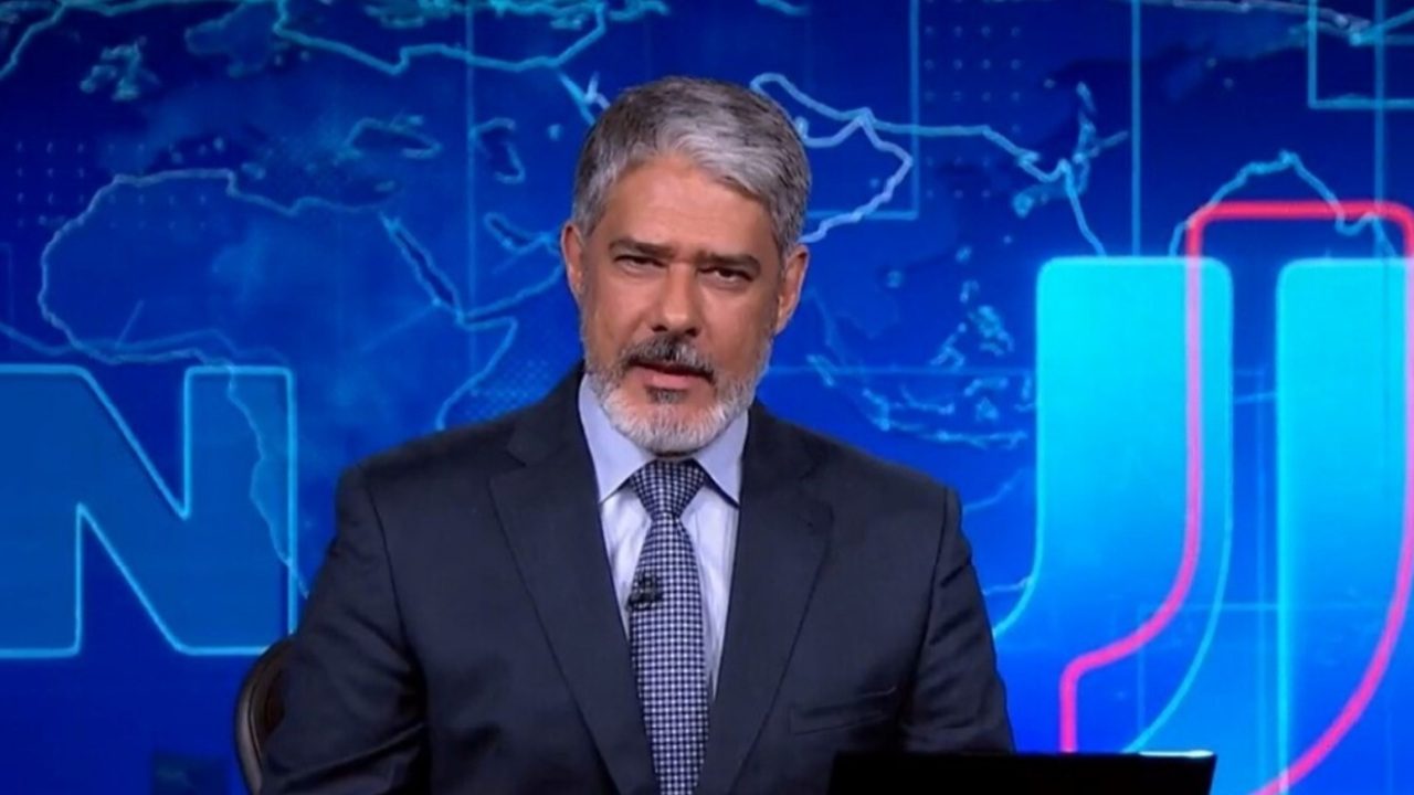 Homem de cabelos grisalhos e barba curta apresenta telejornal em estúdio com fundo azul que exibe mapa-múndi estilizado e letras luminosas, vestindo terno escuro, camisa branca e gravata azul