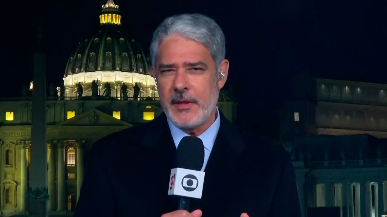 William Bonner com microfone da Globo em frente à Basílica de São Pedro iluminada durante transmissão noturna