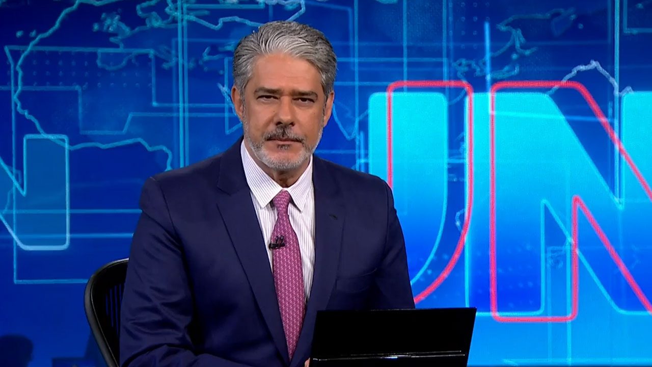 William Bonner de terno azul e gravata rosa apresentando o Jornal Nacional no estúdio da Globo