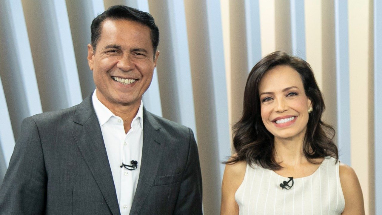 Willian Leite e Fabiana Oliveira sorrindo lado a lado no estúdio do Balanço Geral Manhã com microfones presos à roupa