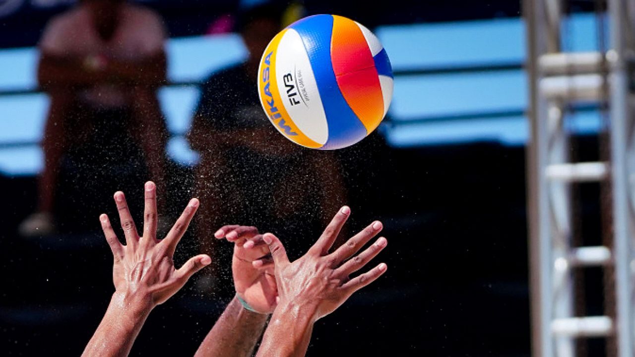 Bola de vôlei Mikasa no ar enquanto jogadores tentam bloqueio em partida de vôlei de praia