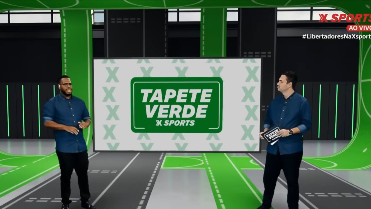 A imagem mostra dois apresentadores em um estúdio esportivo com o logotipo “Tapete Verde X Sports” ao fundo. O cenário é moderno, com tons de verde e cinza remetendo a um campo de futebol. Ambos vestem camisas azul-escuras e conversam durante a transmissão ao vivo, conforme indicado pelo selo “AO VIVO” no canto superior direito