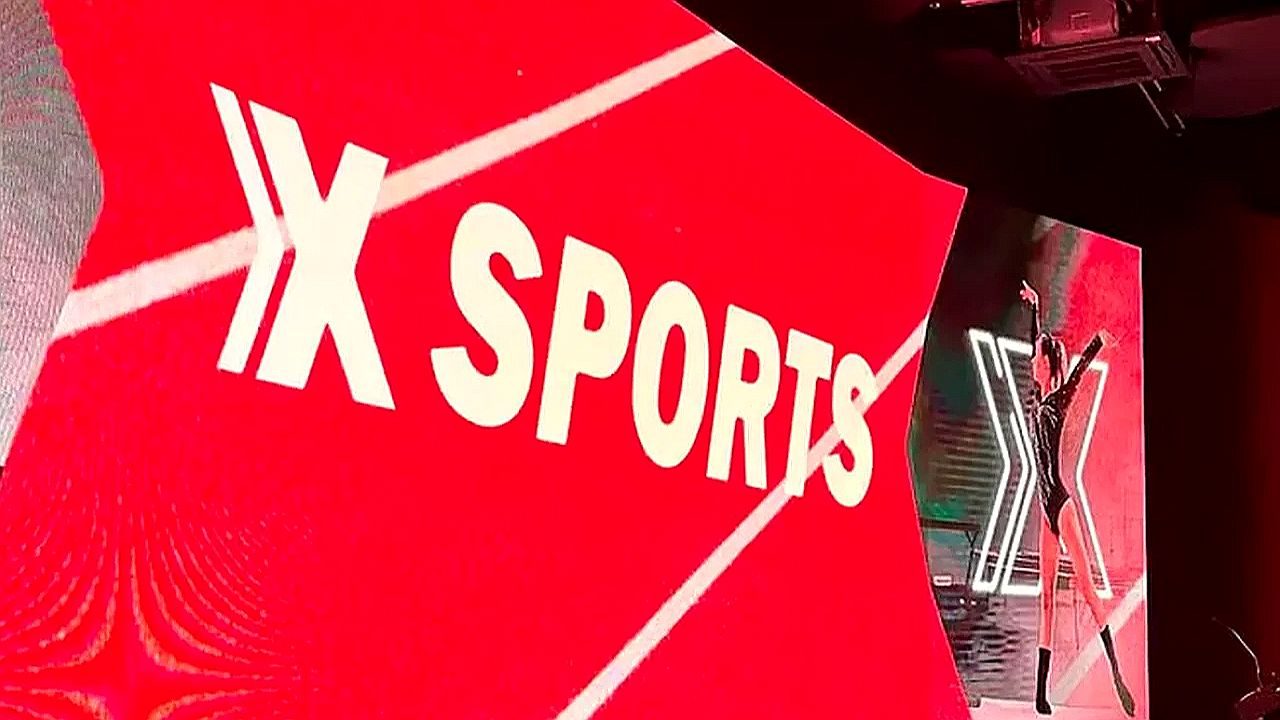 Logotipo do XSports exibido em painel vermelho durante transmissão esportiva em estúdio