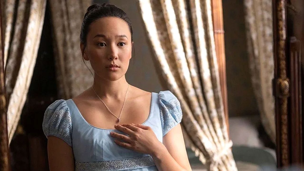 Mulher jovem com vestido de época azul coloca a mão no peito e observa com expressão surpresa em ambiente interno com cortinas ao fundo